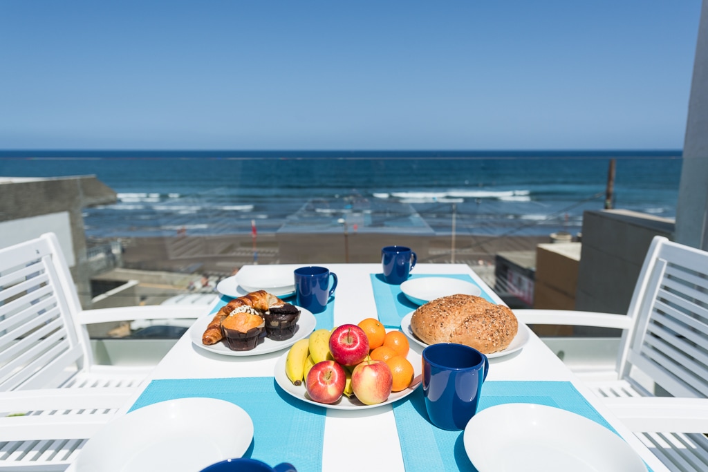 A dining table is set outdoors, overlooking the ocean. A variety of fresh fruits, pastries, and a loaf of bread are arranged on the table, accompanied by blue cups and plates. The expanse of sand and waves is visible in the background under a clear blue sky.