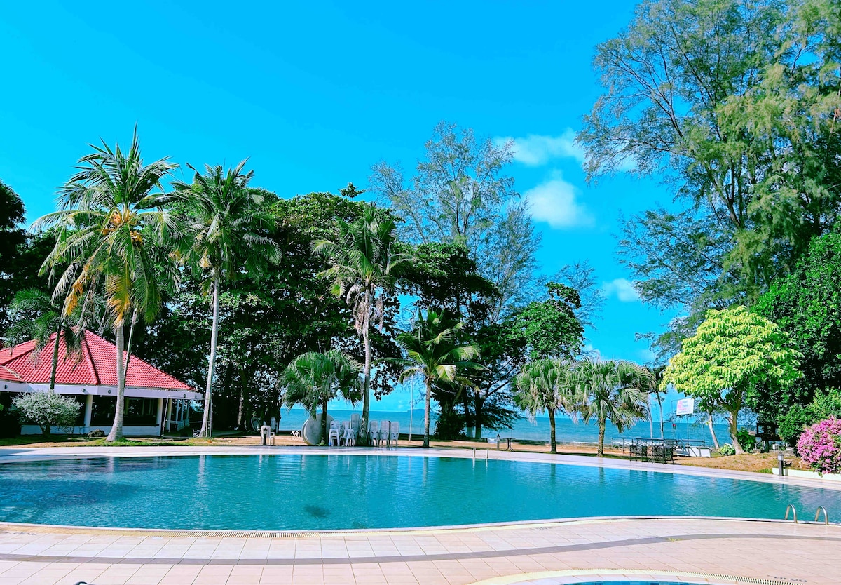 A large swimming pool is surrounded by lush greenery and palm trees. The serene water reflects the clear blue sky and offers views of the nearby beach. A shaded area with tables can be seen in the background, contributing to the tranquil setting.