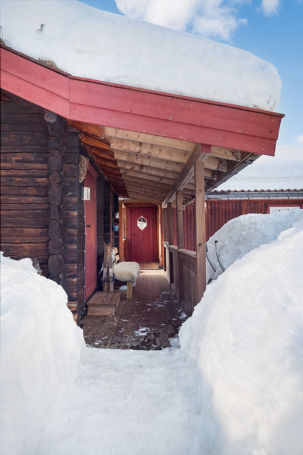 A snow-covered pathway leads to a wooden entrance, flanked by snowdrifts. The rustic cabin is adorned with a red accent on the roof, with a small diamond-shaped window visible on the front door.