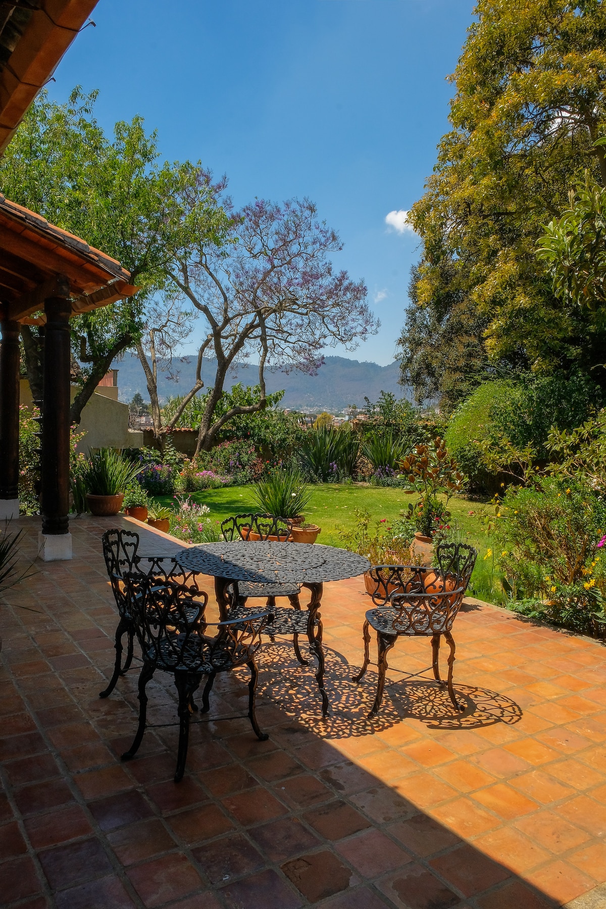A charming outdoor patio features a wrought-iron table surrounded by four intricate chairs. Lush green plants and blooming flowers add color to the private garden, which is framed by tall trees and distant mountains under a clear blue sky.