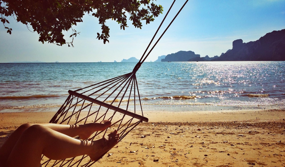 A hammock hangs gently from a tree, with the legs of a person resting comfortably inside. The view of the tranquil beach features gentle waves lapping at the shore, with distant islands visible against a clear blue sky, reflecting sparkling daylight on the water.