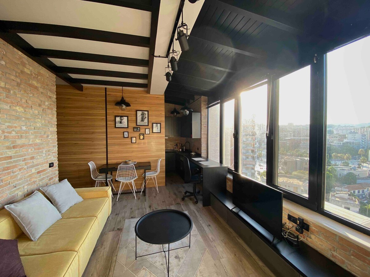A modern living area features a cozy sofa, dining table, and workspace with a view through expansive windows. Exposed brick and wooden accents create a stylish atmosphere, while natural light enhances the space. The area is thoughtfully designed for both relaxation and functionality.