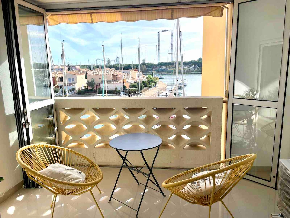 A loggia offers a peaceful seating area with two yellow chairs and a small blue table, overlooking the port. Natural light fills the space, with views of boats and buildings beyond. The balcony is enclosed by sliding glass doors, creating a bright and airy environment.