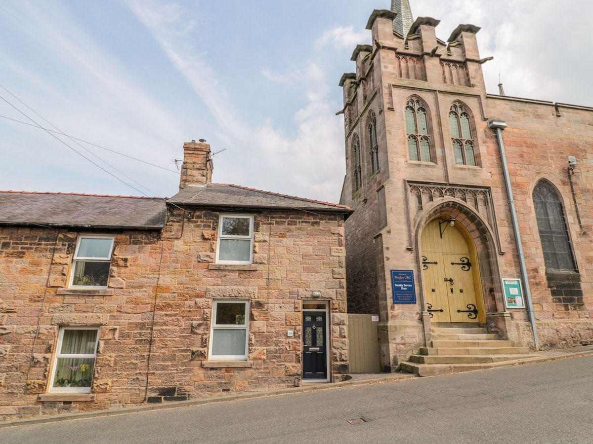 A charming terraced cottage is situated adjacent to a historic stone building. The cottage features a textured stone facade with large windows, while the neighboring structure showcases grand architectural details and arched doorways. The scene captures a peaceful street setting with a gentle slope.