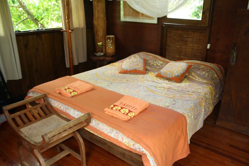 A cozy sleeping area is highlighted by a spacious bed covered with a patterned duvet and topped with two folded towels. Natural wood elements and warm tones contribute to a serene atmosphere. A wooden chair sits beside the bed, complemented by soft natural light streaming through the window.