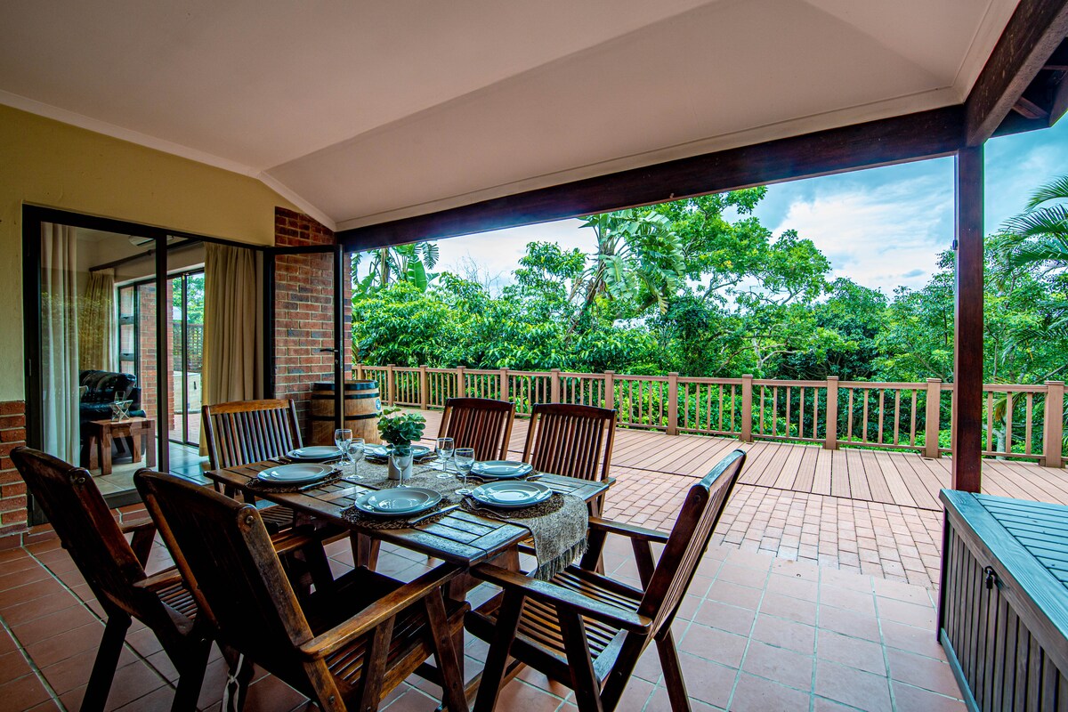 An outdoor dining space features a wooden table surrounded by six chairs. The area is sheltered and opens to a spacious deck, offering views of lush greenery. Natural light filters in through large sliding glass doors, enhancing the connection between indoor and outdoor areas.