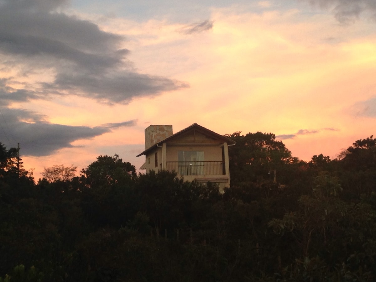 The house is positioned among lush greenery, showcasing a warm architectural style against a vibrant sunset sky. The exterior features a balcony that offers a view of the surrounding landscape, creating a serene connection with nature.