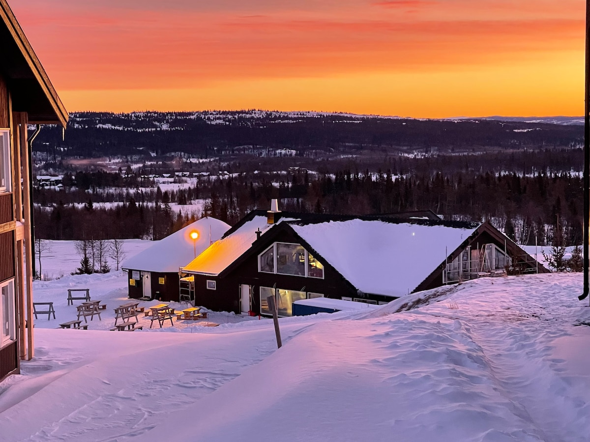 A snow-covered landscape is illuminated by a soft sunset, casting warm hues over a cluster of ski lodges. Outdoor seating areas are visible, set against the backdrop of distant hills and a serene valley. The scene invites tranquility and outdoor activities.