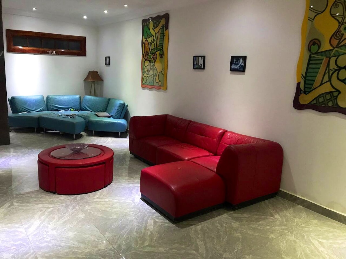 A spacious living area features a vibrant red sectional sofa positioned around a low, circular table. Soft blue couches are arranged in the background, complemented by artwork adorning the walls. Ambient lighting enhances the contemporary decor, creating a relaxed atmosphere.
