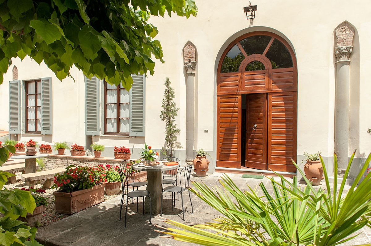 An entrance features a large wooden door framed by stone columns, inviting guests into the property. Surrounding greenery and flower pots create a welcoming atmosphere in the courtyard. A round table and chairs are set in the outdoor area, enhancing the inviting space.