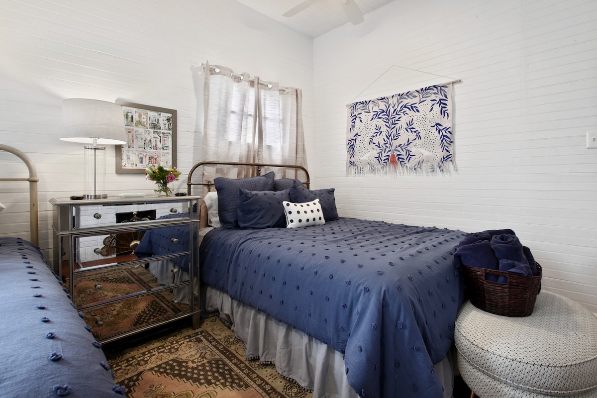 A serene bedroom is presented, featuring two beds dressed in deep blue bedding. Light-colored walls complement the space, while natural light filters through sheer curtains. A wicker basket and decorative pillow add texture, and a wall hanging with a botanical design enhances the room's aesthetic.