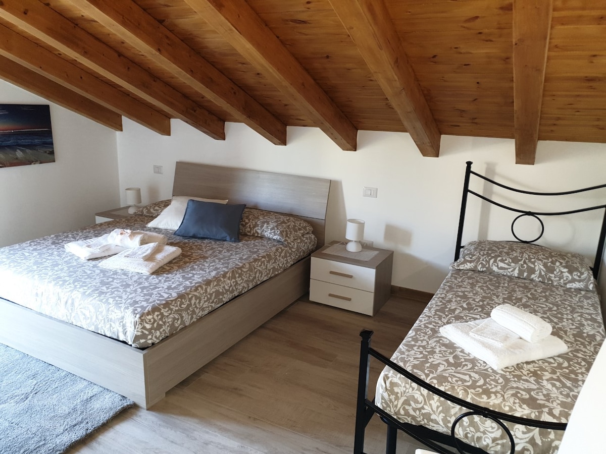 The room features a spacious bed and a single bed, both adorned with light bedding. Wooden beams accent the ceiling, enhancing the airy feel. A bedside table with lamps is positioned next to the larger bed, while a soft rug lies on the wooden floor.