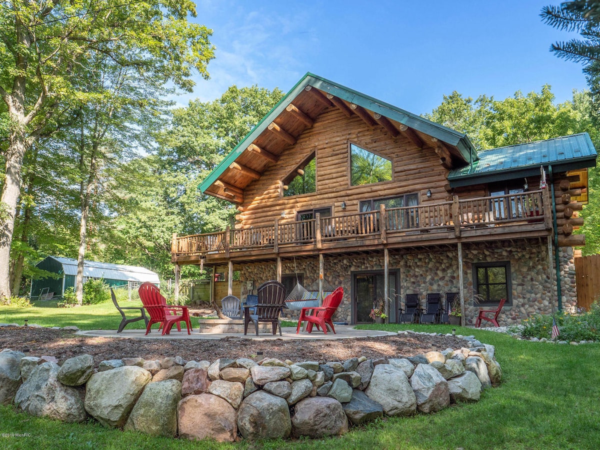 A spacious log cabin features a combination of stone and wood detailing. A large deck extends across the upper level, complemented by multiple outdoor seating areas arranged around a circular rock fire pit. Lush greenery surrounds the cabin, enhancing a serene outdoor setting.