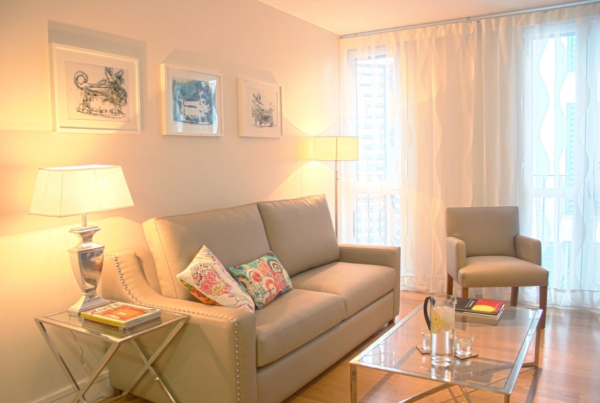 A light-filled living area is furnished with a soft beige sofa and a decorative armchair. A glass coffee table holds art books and candles, while framed artwork hangs on the wall. Sheer curtains allow natural light to softly illuminate the space.