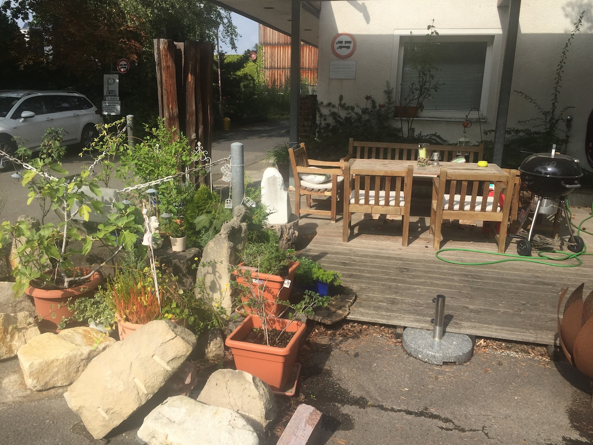 An outdoor seating area features wooden benches and a table, surrounded by various potted plants and greenery. Natural stone elements are placed throughout the space, enhancing the garden-like atmosphere. A grill is positioned nearby, with a concrete surface visible, contributing to the functionality of the area.
