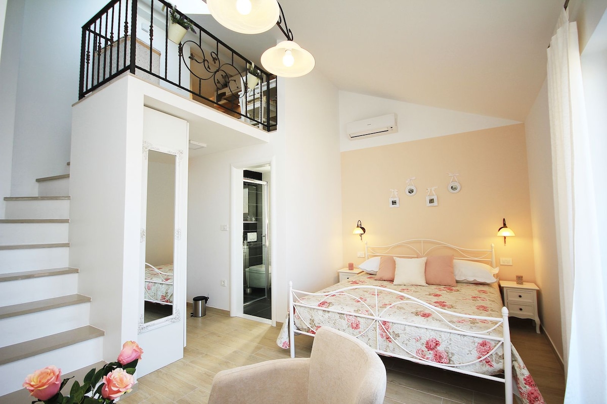 An air-conditioned room features a spacious bed adorned with floral bedding. A cozy sitting corner is present, along with a staircase leading to a gallery. The room is complemented by two wall-mounted lights and a mirrored wardrobe, offering functionality and style.