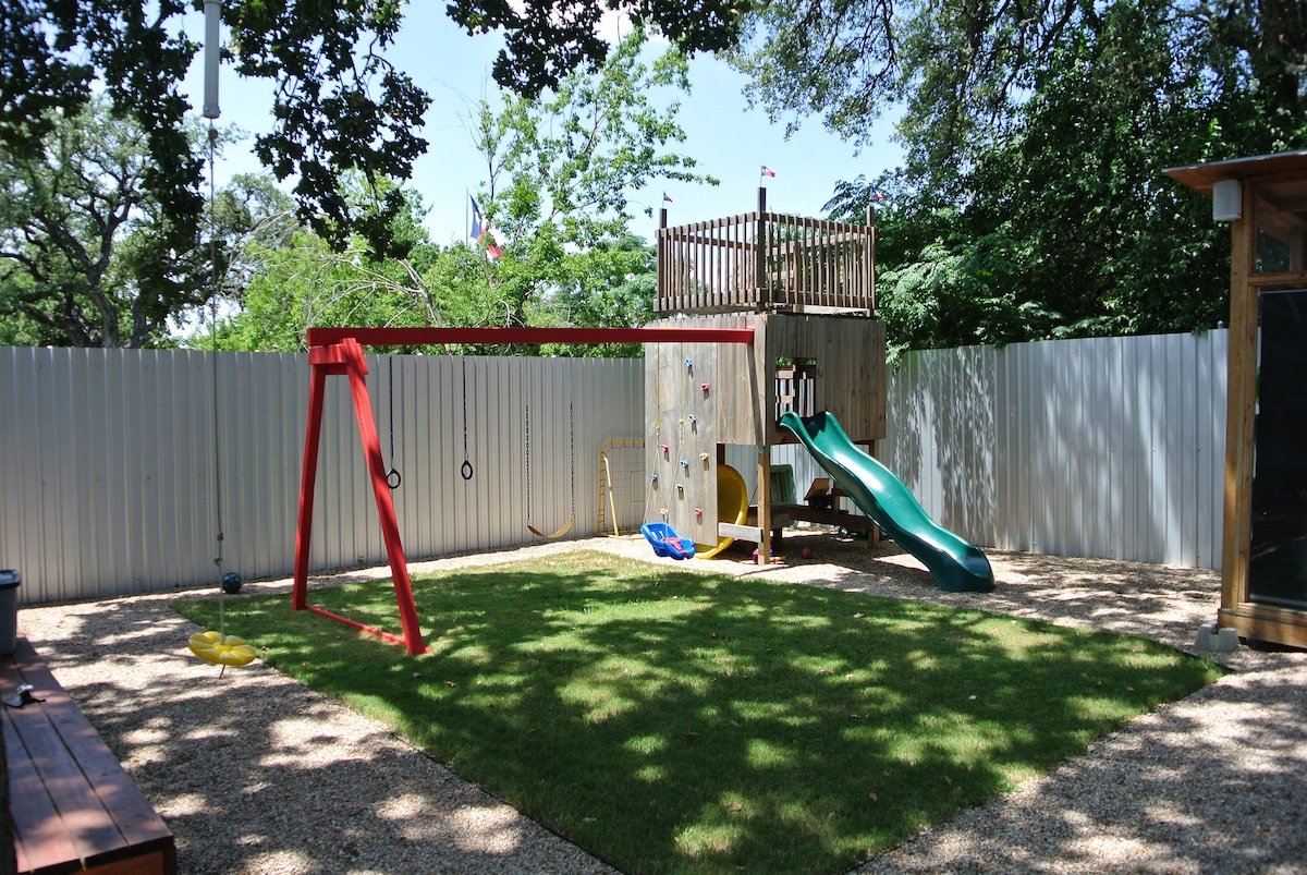 A spacious outdoor play area features a green lawn bordered by a white fence. A red swing set and a wooden fort with a slide are visible, along with bright climbing equipment. Sunlight dapples the area through surrounding trees, creating a cheerful play environment.