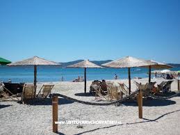 A serene beach scene presents several sun loungers beneath straw umbrellas, positioned on soft, white sand. Calm blue waters stretch in the background, with a clear sky overhead. A hammock is gently swaying between two posts, offering a relaxing spot for visitors.