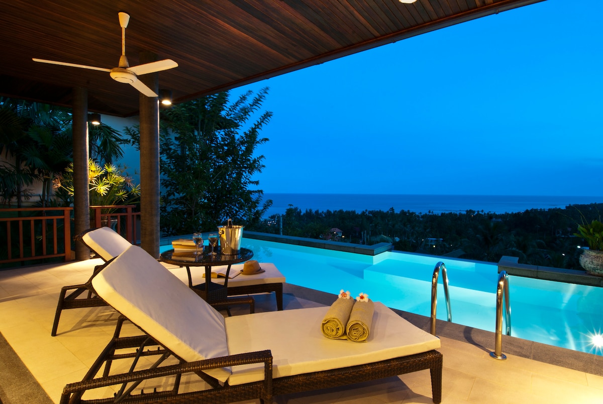 A sunbathing terrace is featured, equipped with lounge chairs and a small table. A refreshing infinity pool extends into the horizon, offering scenic views of the landscape and ocean. Soft lighting enhances the calm atmosphere as dusk settles in.