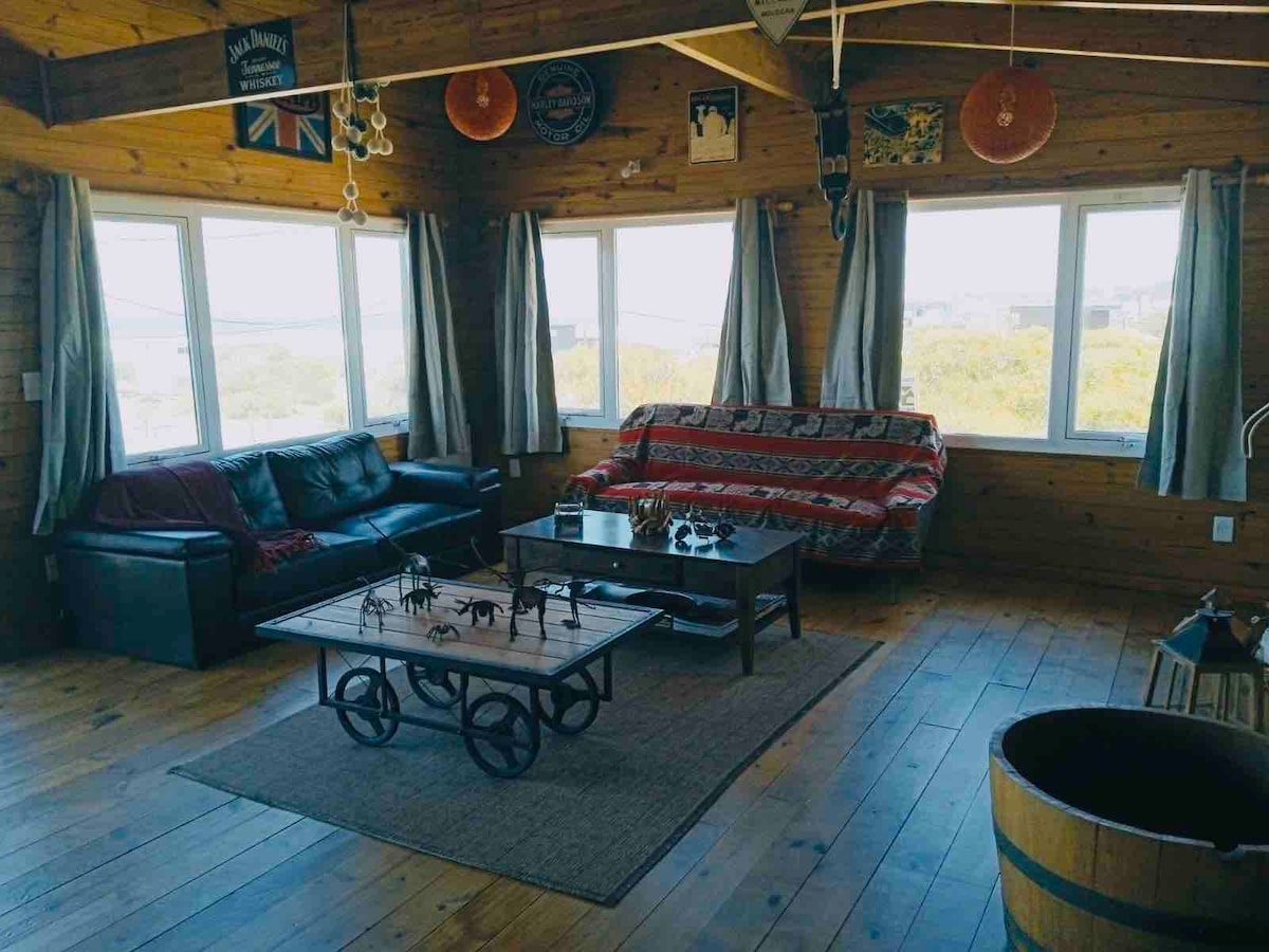 A spacious living area features large windows that allow natural light to fill the room. Two comfortable sofas are arranged around a wooden coffee table with decorative items. A warm wooden floor enhances the cozy ambiance, complemented by soft curtains and charming wall decor.