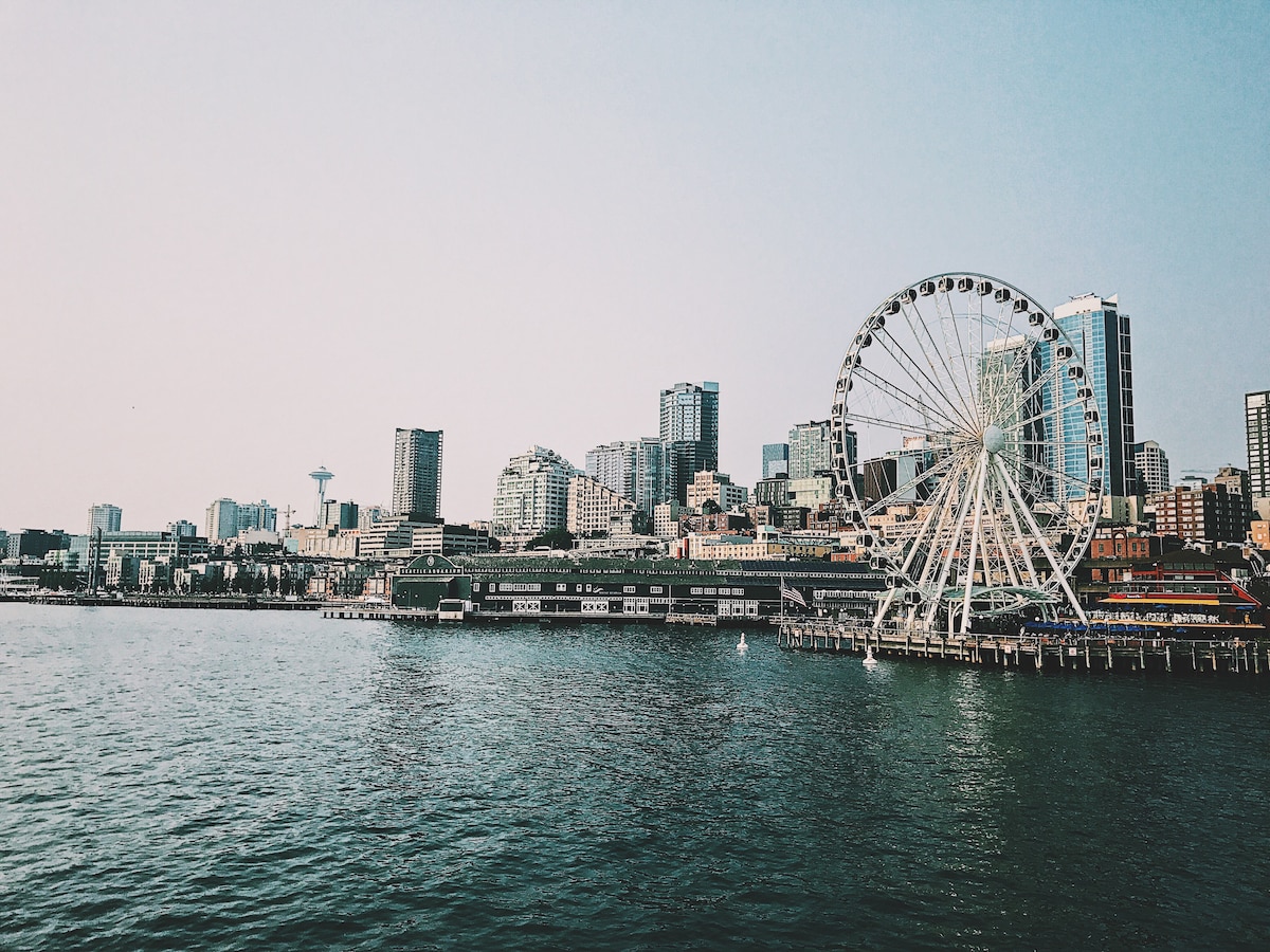 A panoramic view of the Seattle waterfront is presented, showcasing the iconic Seattle Great Wheel alongside the city's skyline. Various buildings rise in the background, with the Space Needle visible among them. The calm waters reflect the structures, enhancing the city's coastal charm.