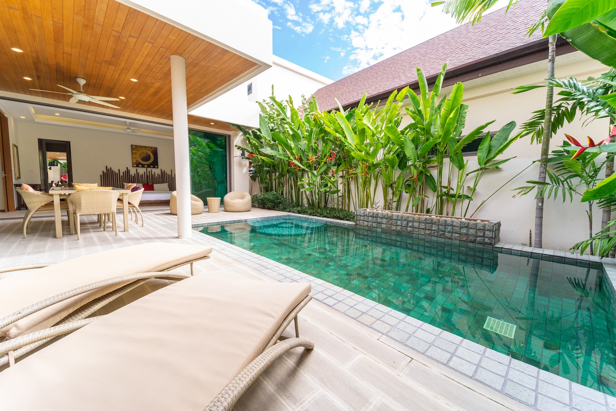 A tranquil outdoor area features a private pool surrounded by lush greenery. Comfortable lounge chairs are positioned near the water, enhancing the relaxation space. A covered terrace is visible in the background, providing a shaded area for dining and leisure.