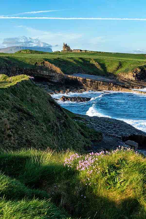 A coastal view captures lush green cliffs meeting the ocean, with gentle waves lapping against the rocky shore. A historic structure is situated atop the hill, overseeing the landscape. Vibrant wildflowers add a natural touch to the foreground, enhancing the serene setting.