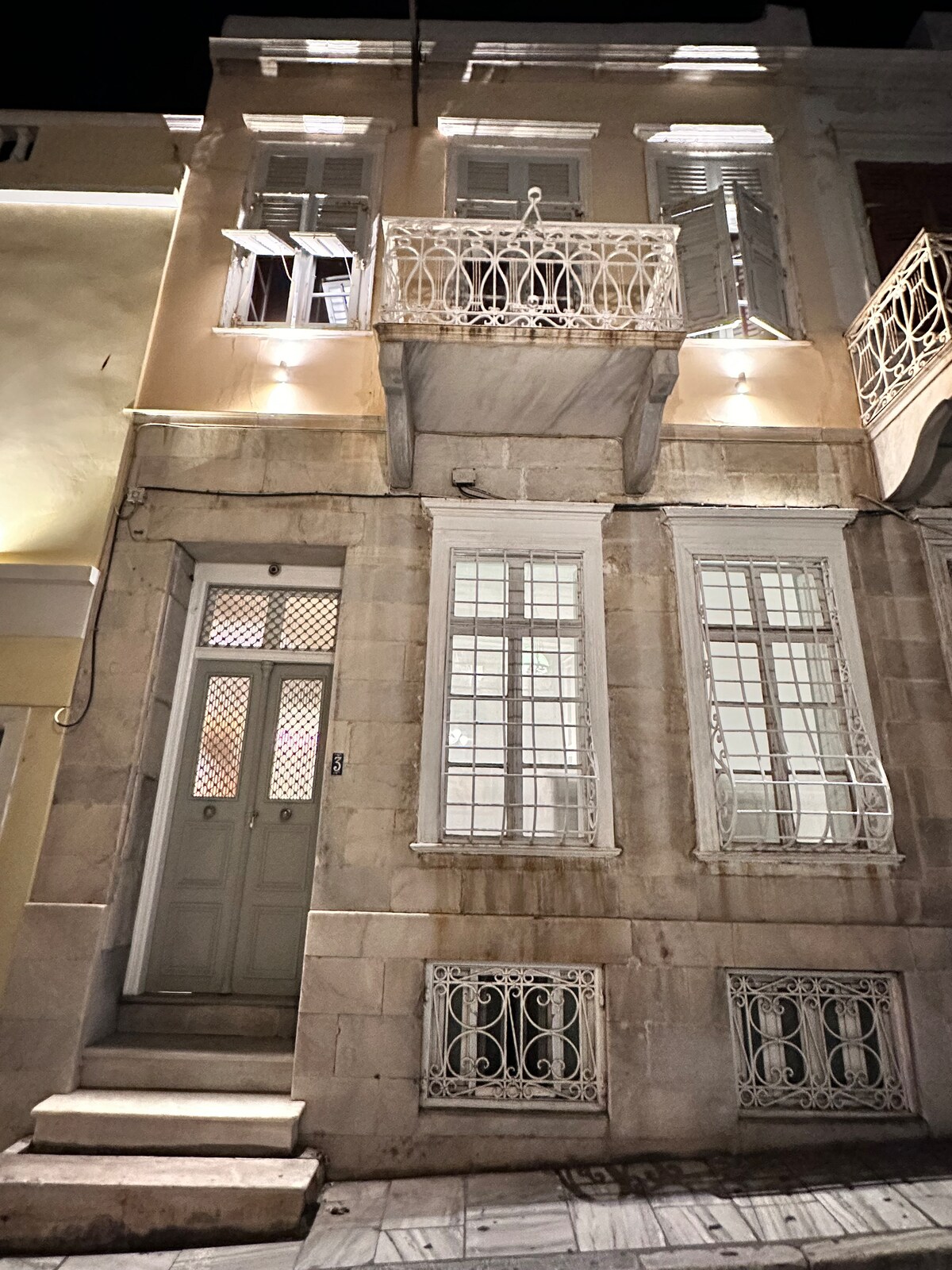 The neoclassical facade of the mansion features intricate ironwork balconies and large windows with shutters. Soft lighting highlights the stonework, enhancing the building's elegance at night. A set of steps leads to the main entrance, framed by decorative elements and a welcoming atmosphere.