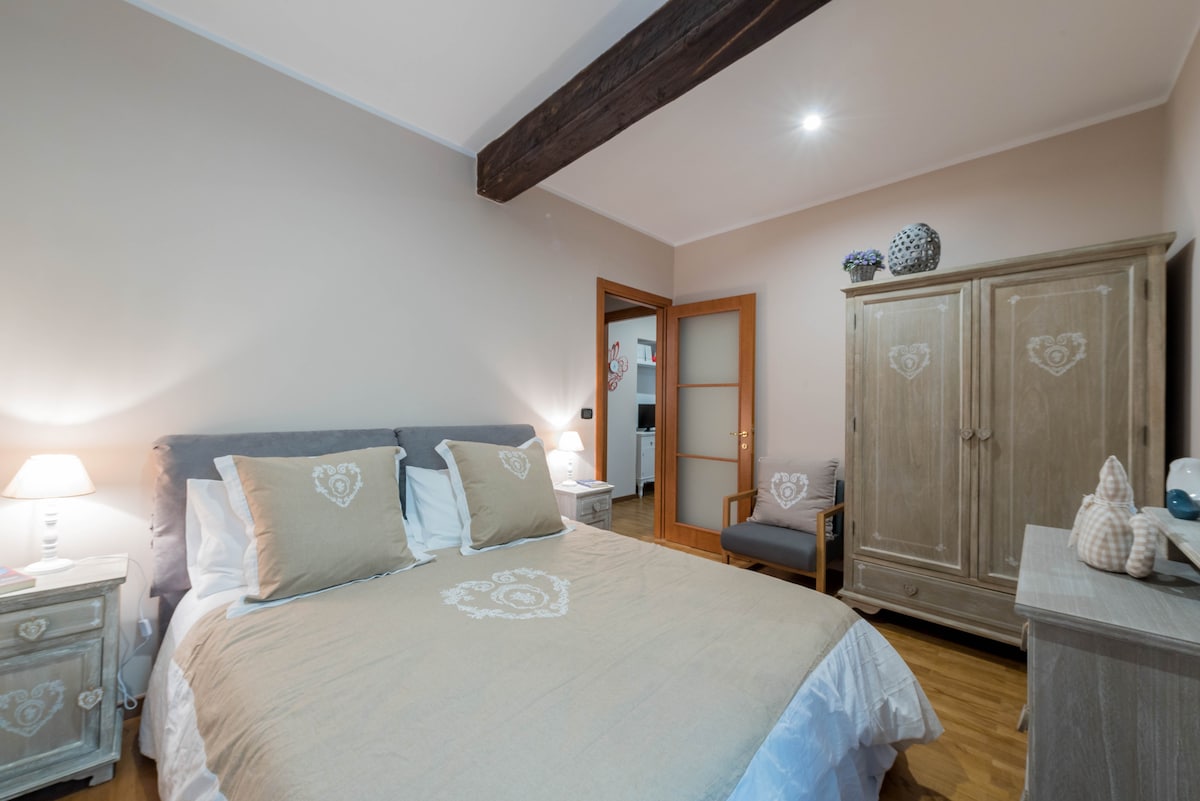 A cozy bedroom features a spacious bed with light-colored linens and decorative pillows. Nightstands with lamps are on either side of the bed. A wooden wardrobe and a chair stand against the wall, with soft lighting enhancing the calm atmosphere.