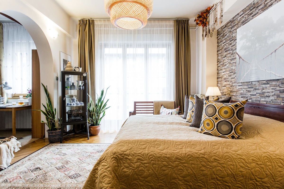A spacious living area is highlighted by a large bed adorned with a textured blanket and decorative pillows. A warm light fixture hangs above, while plants and a cabinet add touches of greenery. Natural light floods in through multiple large windows, enhancing the room's inviting ambiance.