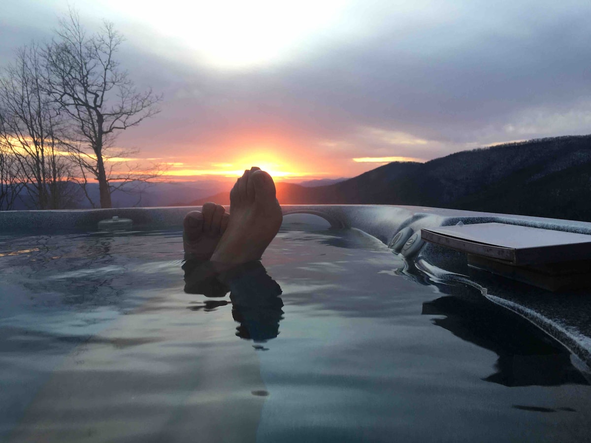 The image captures a tranquil moment in a hot tub, with feet visible above the water's surface. A vibrant sunset casts warm hues over the mountains in the background, while trees frame the serene landscape, creating a sense of relaxation and connection with nature.