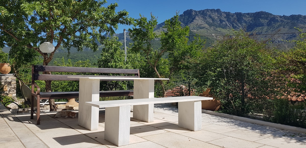A simple outdoor seating area features a wooden bench and white stone tables. Surrounding greenery includes trees and shrubs, set against a backdrop of mountains. Clear blue skies contribute to a sense of openness and tranquility in the space.