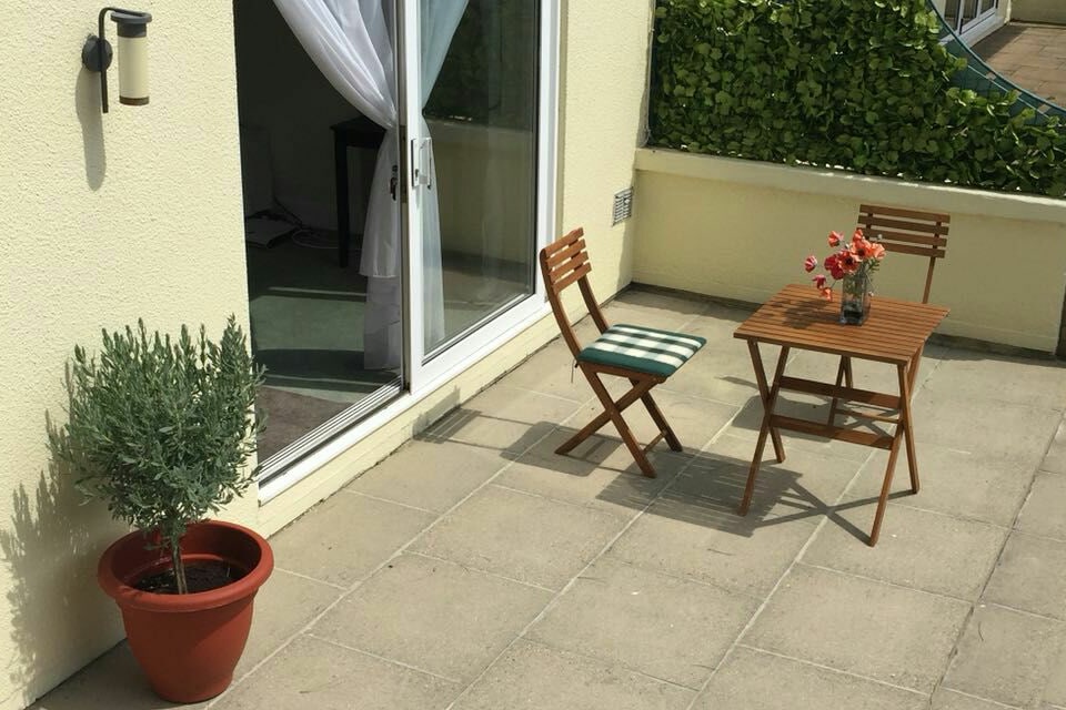 A private patio area features a small wooden table and two folding chairs, complemented by a potted plant. A simple vase with fresh flowers sits on the table. The patio overlooks a well-maintained garden hedge, providing a serene space for relaxing.