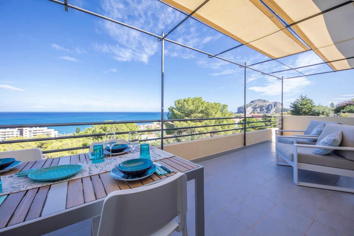 A spacious terrace offers a view of the sea and surrounding greenery. A wooden dining table is set with turquoise dishware and glasses, complemented by white chairs. Lounge seating in the corner invites relaxation while enjoying the panoramic scenery.