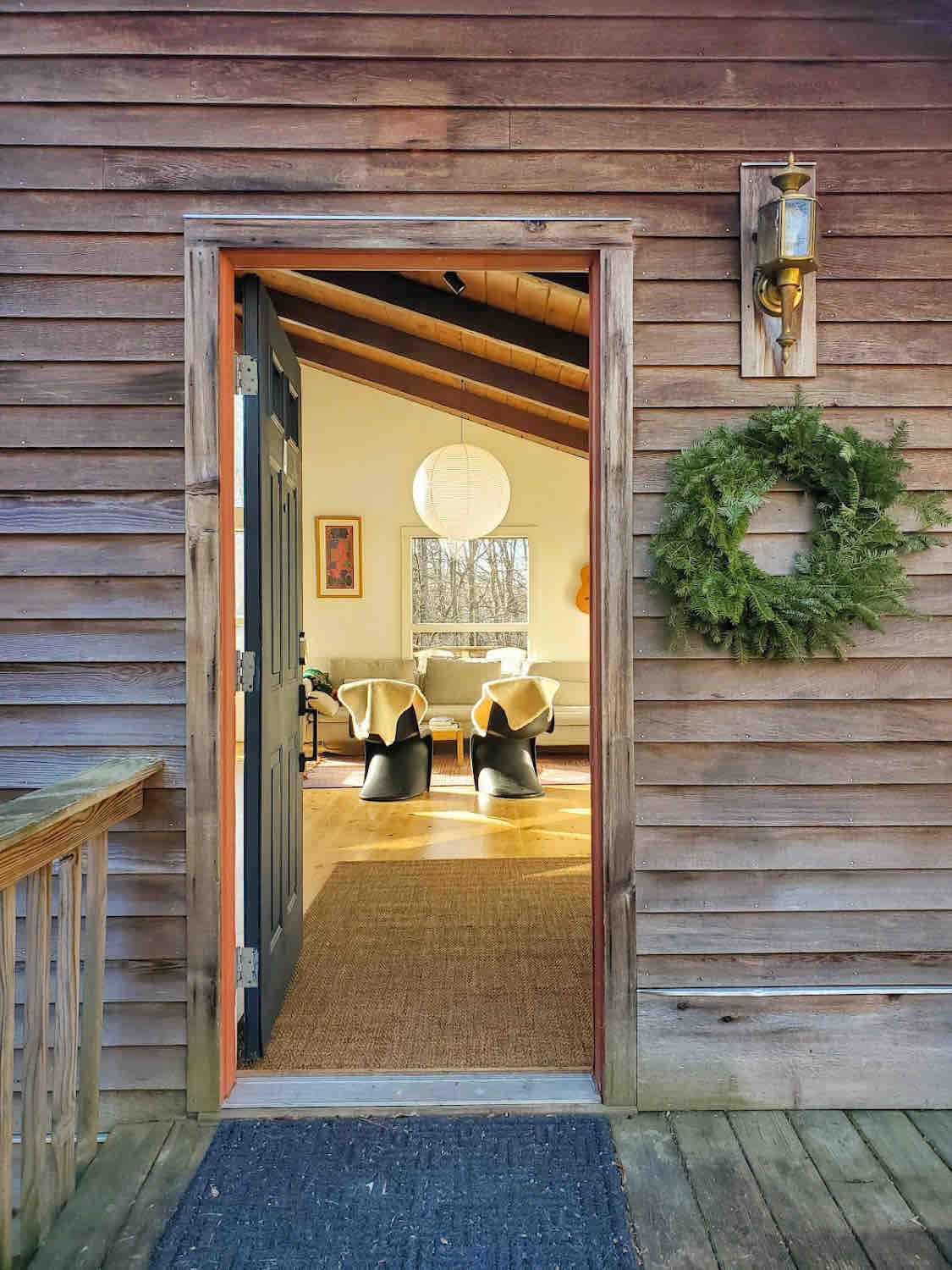 The entryway features a welcoming entrance with a wooden door, adorned with a green wreath. A glimpse into the bright interior reveals modern seating and natural light streaming through large windows, enhancing the inviting space that awaits inside.