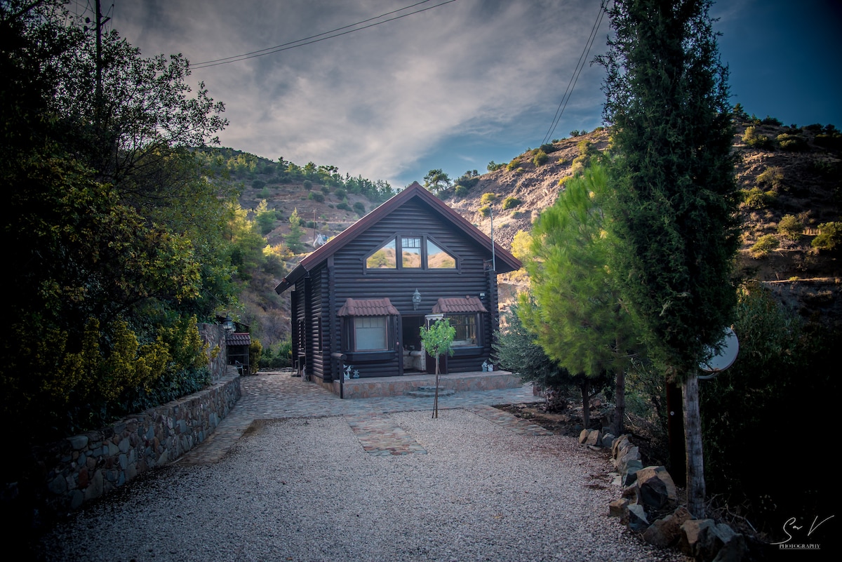 A log house is positioned amidst earthy tones and greenery, framed by rugged hills. The structure features a peaked roof with large windows reflecting natural light, enhancing the wooden exterior. A gravel path leads to the entrance, surrounded by shrubs and trees.