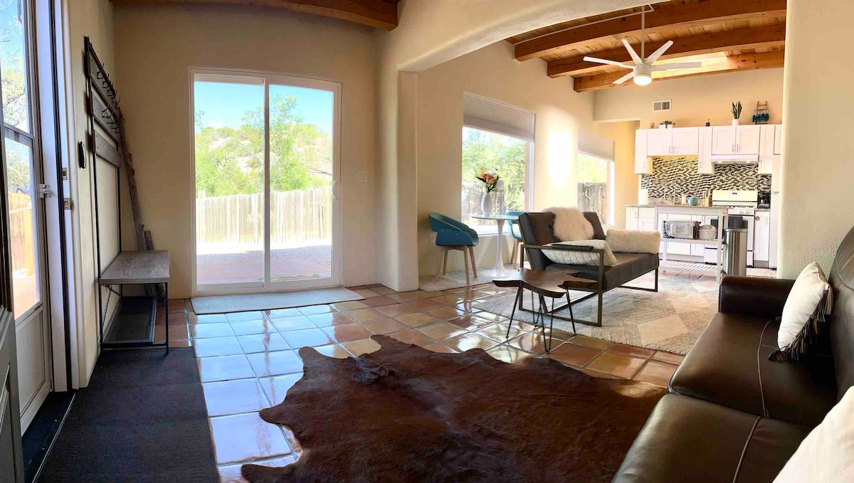 A spacious living area is showcased, featuring a leather sofa and a small coffee table atop a textured rug. Large windows allow natural light, and an adjacent open kitchen is visible. The room includes high wood-beamed ceilings and a modern ceiling fan.