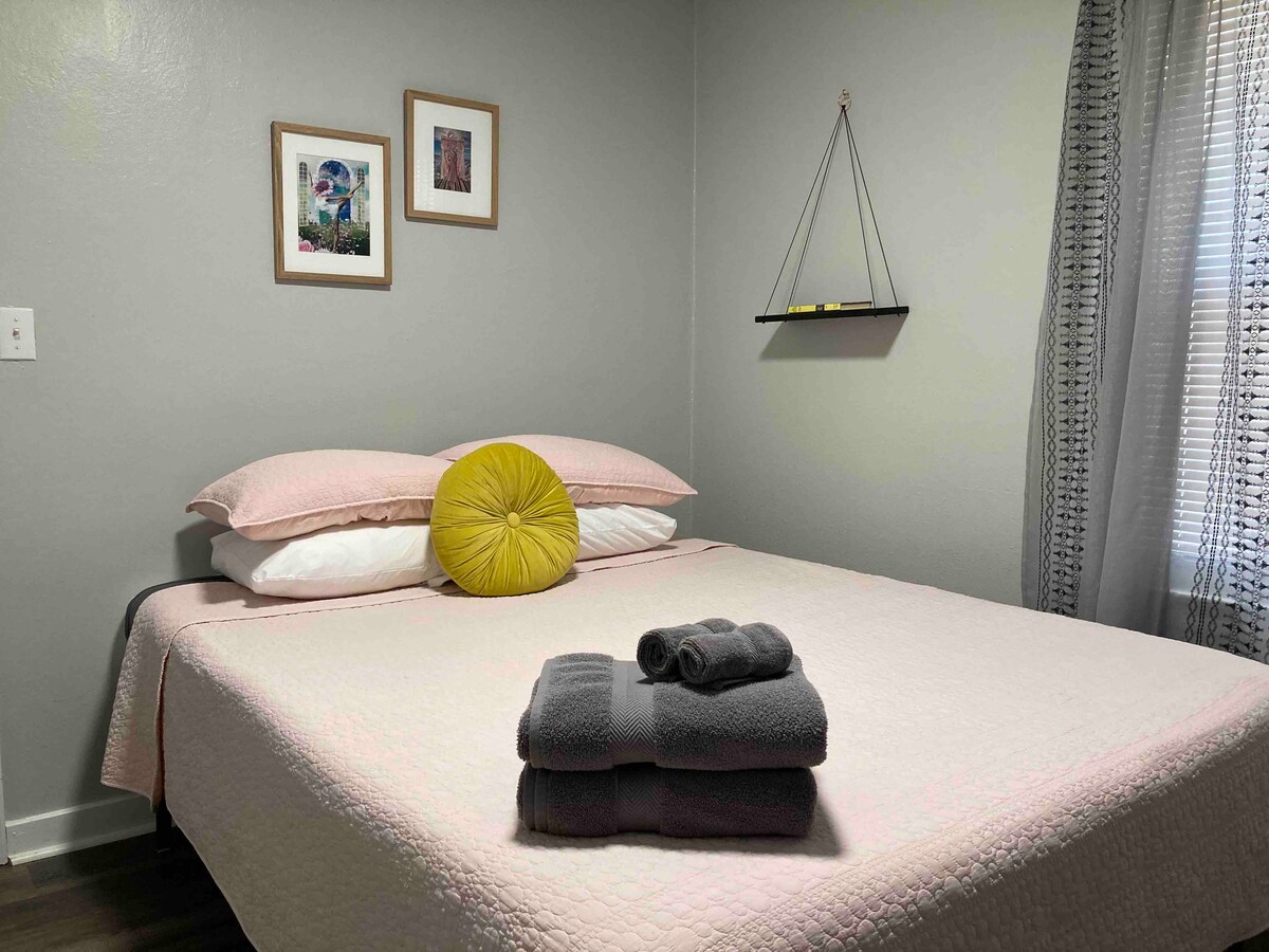 A cozy sleeping area features a spacious bed with a light pink quilt, layered with white pillows and a yellow accent pillow. Soft towels are neatly arranged at the foot of the bed. Natural light enters through a window, complemented by flowing curtains.