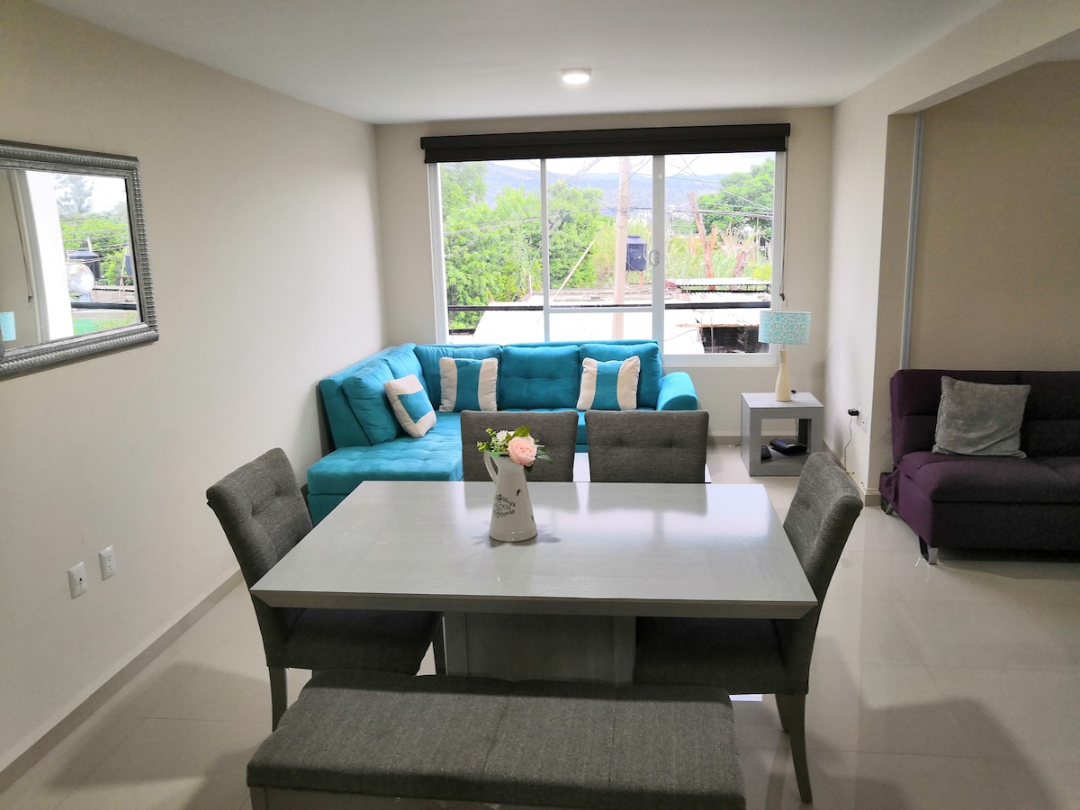 A modern living area features a light-colored table accompanied by gray upholstered chairs. A turquoise sectional sofa is positioned against the wall, beside a purple couch. Large windows allow natural light to illuminate the space and provide a view of the surrounding greenery.