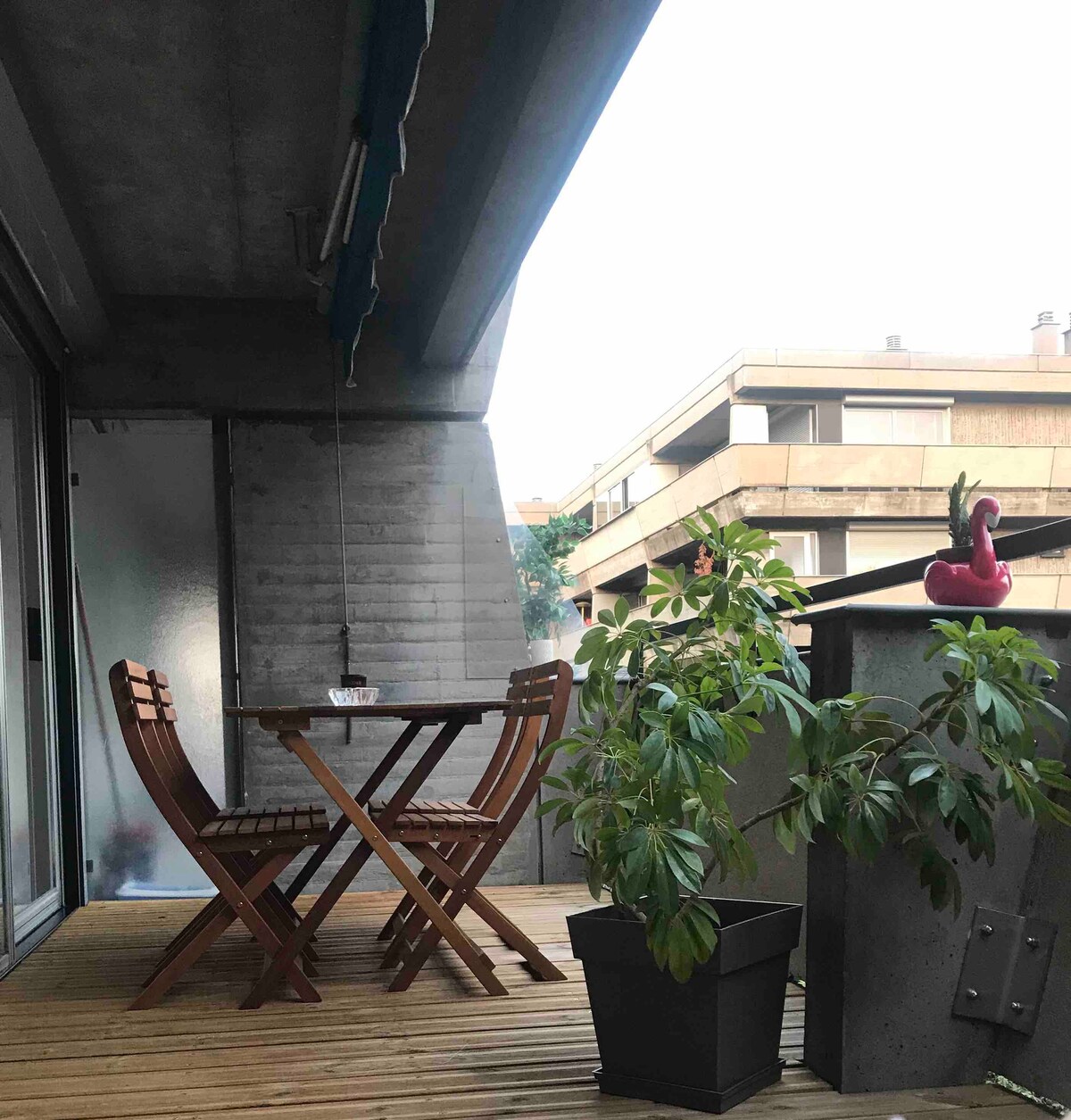 A private terrace features a small wooden table and two folding chairs, framed by potted plants. The balcony provides a view of the surrounding buildings, with a shaded area created by an awning overhead.