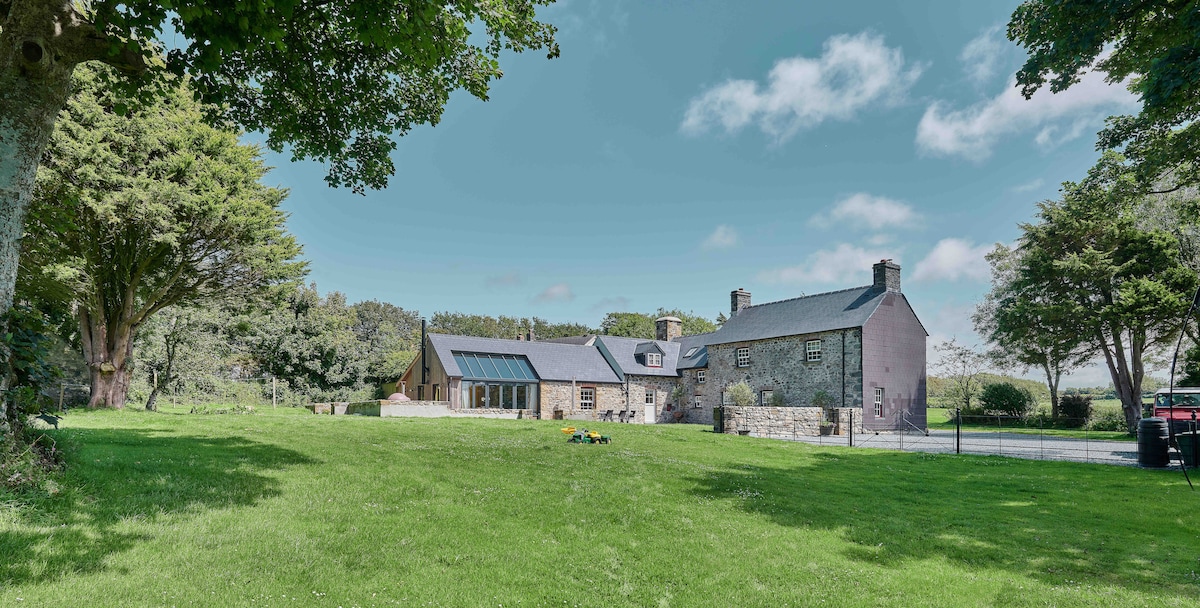 A traditional Welsh farmhouse is surrounded by lush green lawns and mature trees. The building features a blend of stone and timber with large windows reflecting the sky. The extensive grounds provide an open space for relaxation, contributing to a serene countryside setting.