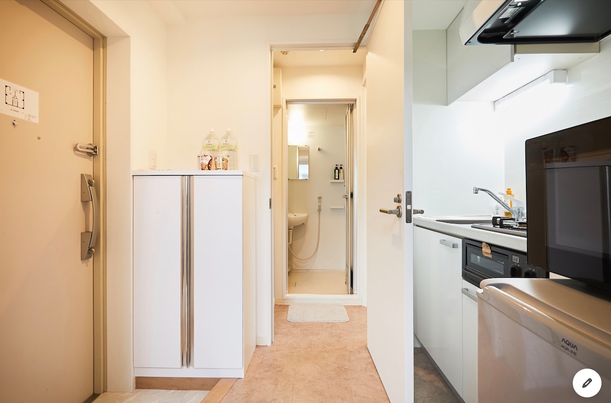 The entrance showcases a compact hallway leading to a well-equipped kitchen and a bathroom. A white refrigerator stands near the door, while modern appliances, including a stove and washing machine, are visible, contributing to a functional living space.