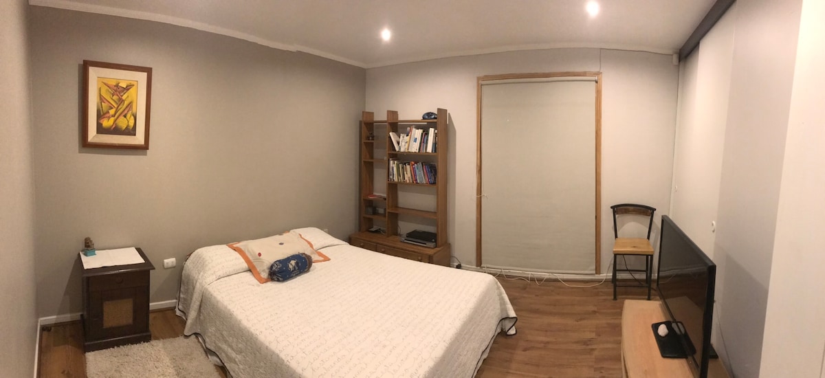 A spacious bedroom features a large bed with a white blanket and decorative pillow. A bookshelf filled with books is positioned beside the bed, while a wooden chair and a television stand are visible across the room. Natural light filters through a window covered by a sliding panel.