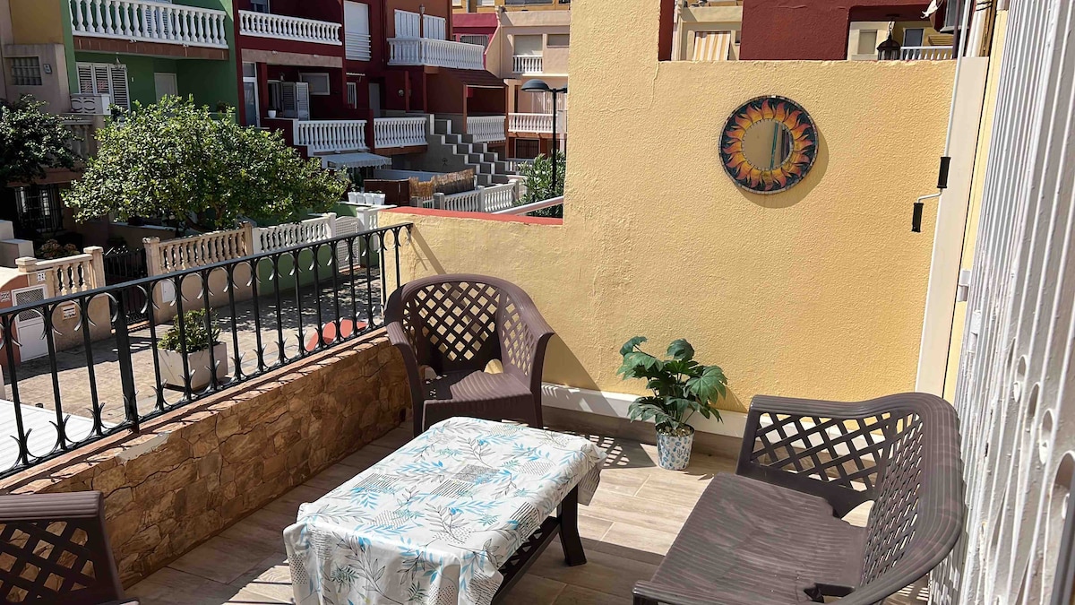 An outdoor terrace is featured, furnished with a table and two armchairs. A small potted plant is positioned nearby. The terrace overlooks a communal area, with homes in the background displaying colorful facades and balconies.