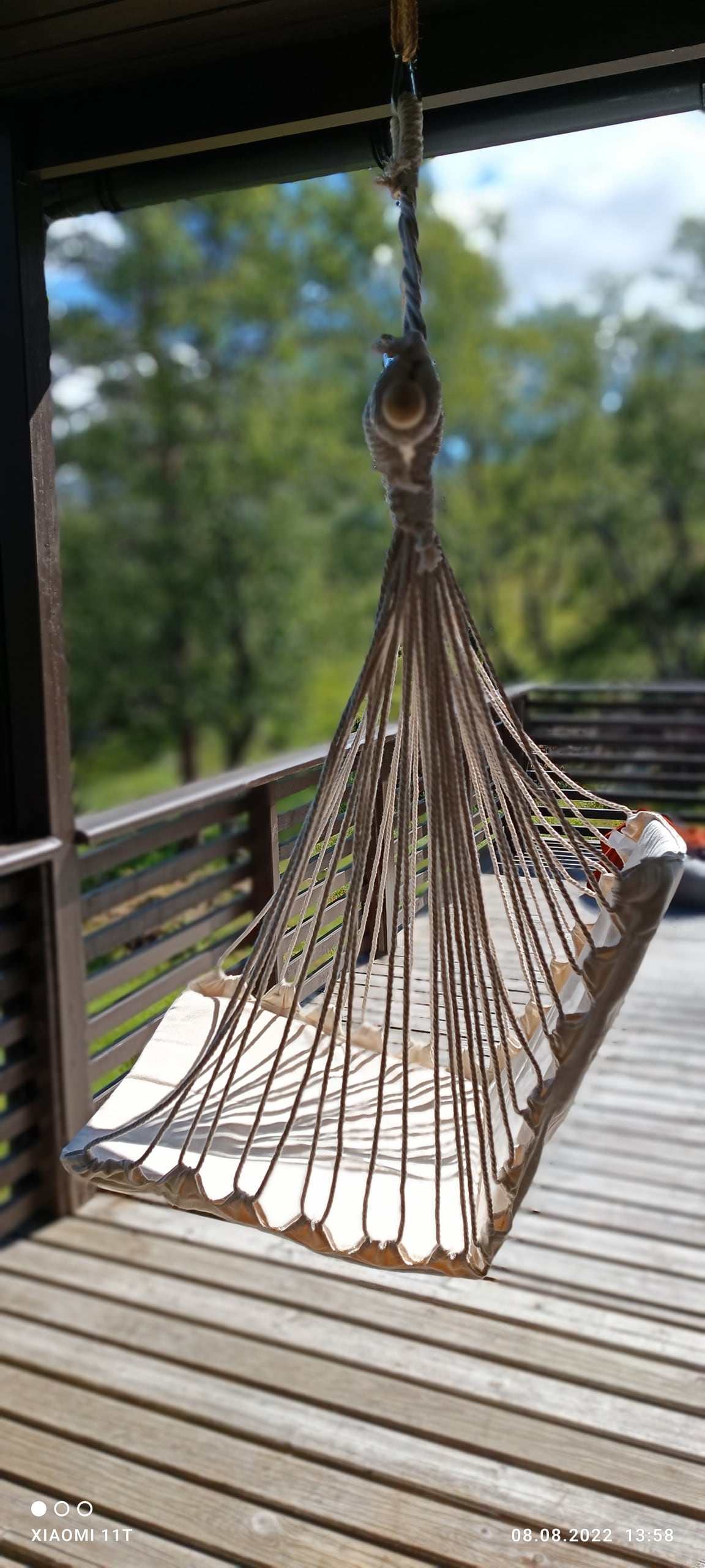 A hanging hammock made of light-colored fabric and natural fibers is anchored from a wooden porch. The surrounding outdoor area features a wooden deck and green foliage in the background, adding a serene atmosphere for relaxation.