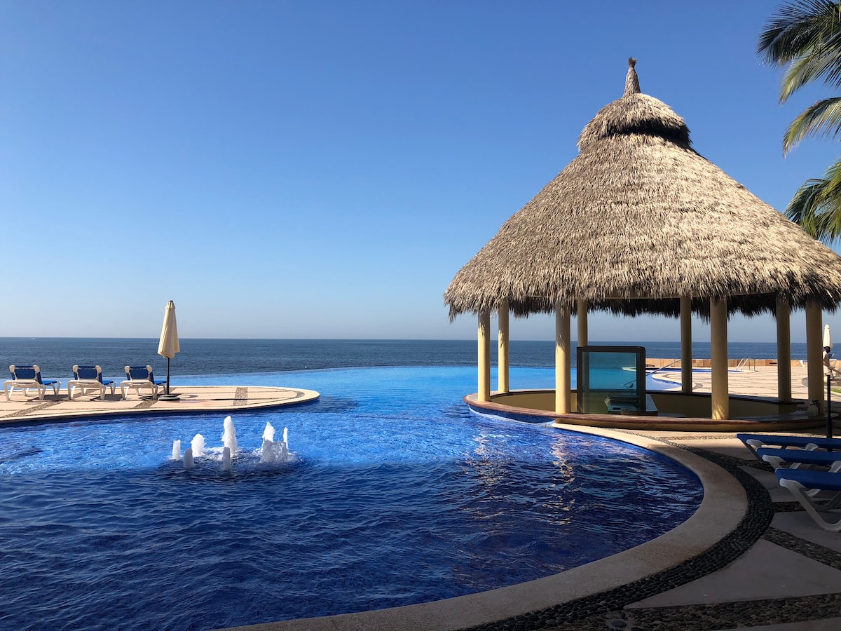 An expansive outdoor pool is depicted with a straw-roofed palapa overlooking the ocean. A fountain gently bubbles in the center of the pool, and lounge chairs are arranged nearby, inviting guests to enjoy the serene water views under clear blue skies.
