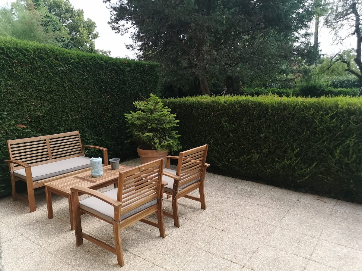 An outdoor seating area is visible, featuring two wooden chairs and a matching bench with light-colored cushions. A table is positioned at the center. Lush green hedges provide privacy, while a potted plant adds a touch of nature to the setting.