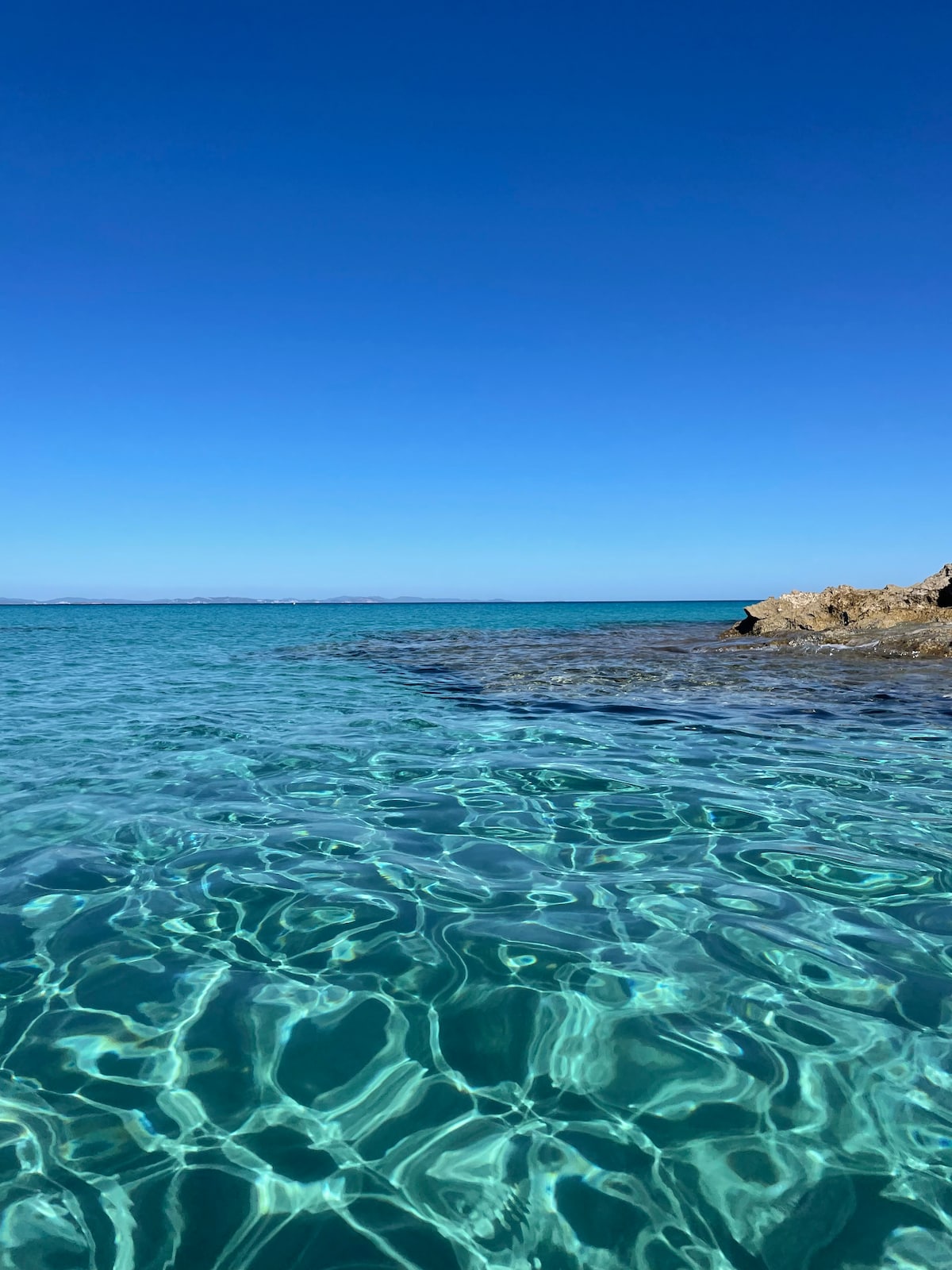 The image captures a clear turquoise sea under a bright blue sky. The water is calm, displaying a rippling surface that reflects sunlight, with rocky formations visible in the foreground, enhancing the natural beauty of the coastal scene.