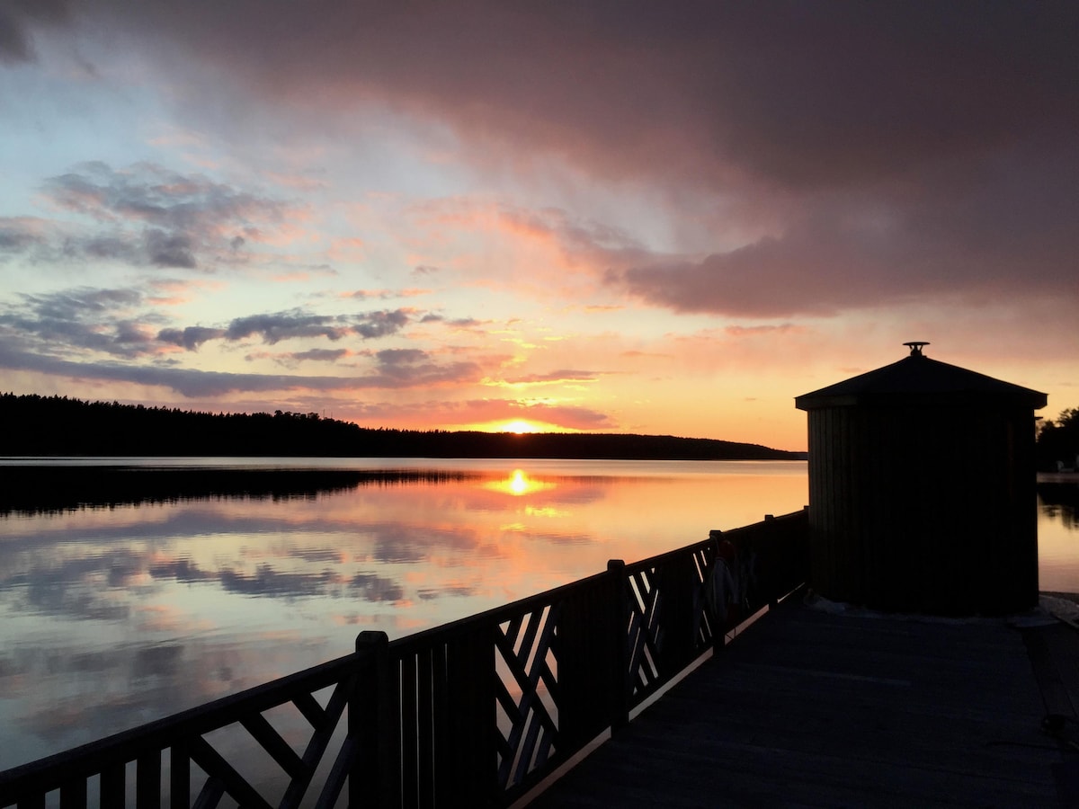 A tranquil sunset over Lake Mälaren is captured, reflecting vibrant hues across the calm water. A wooden dock extends into the lake, framed by a nearby structure that provides shelter. The surrounding woods are softly silhouetted against the colorful sky.