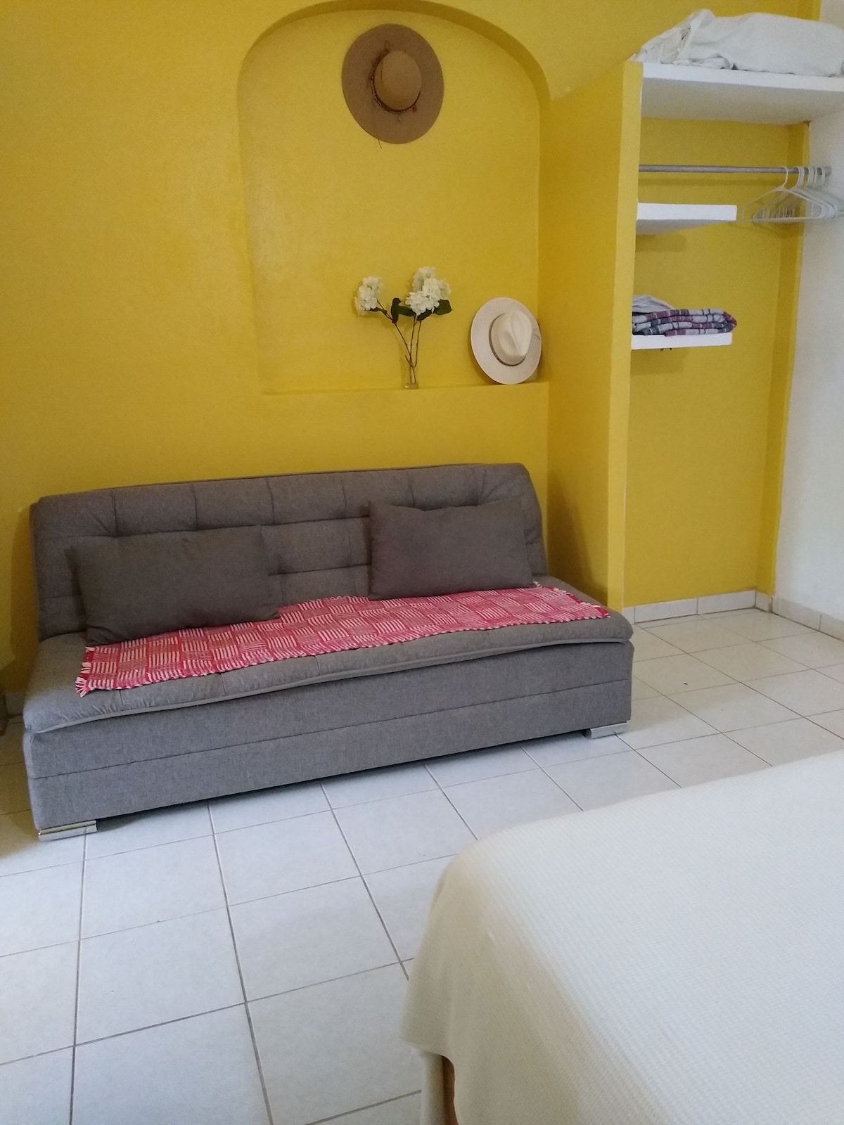 A bright yellow wall creates a warm backdrop for the seating area, featuring a gray sofa with cushions and a woven blanket. A small shelf holds hats and folded items, while a decorative arrangement of flowers adds a touch of character.
